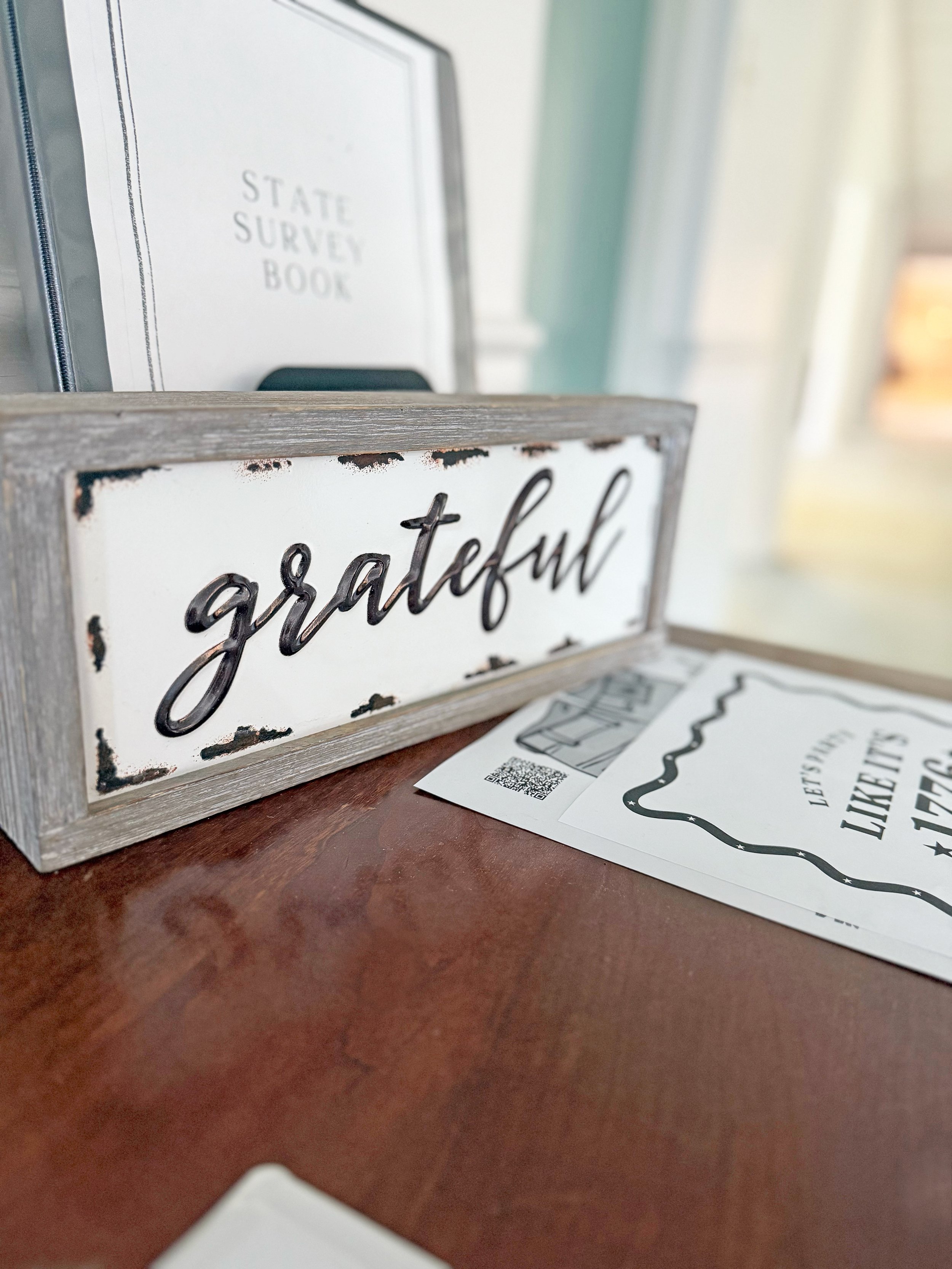 A wooden sign with the word 'grateful' in black script lettering, placed on a wooden table. Behind it, there is a blurred sign that reads 'STATE SURVEY BOOK' and some papers with printed text and QR codes.