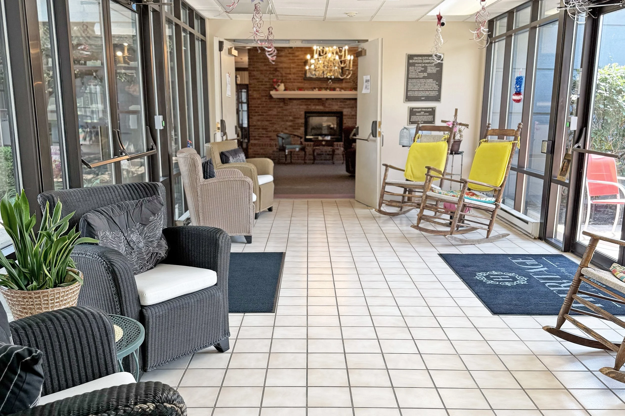 A cozy waiting area with wicker chairs, a plant, and a black and white couch, near glass doors, with rocking chairs with yellow cushions, and a fireplace in the background decorated for the holidays.