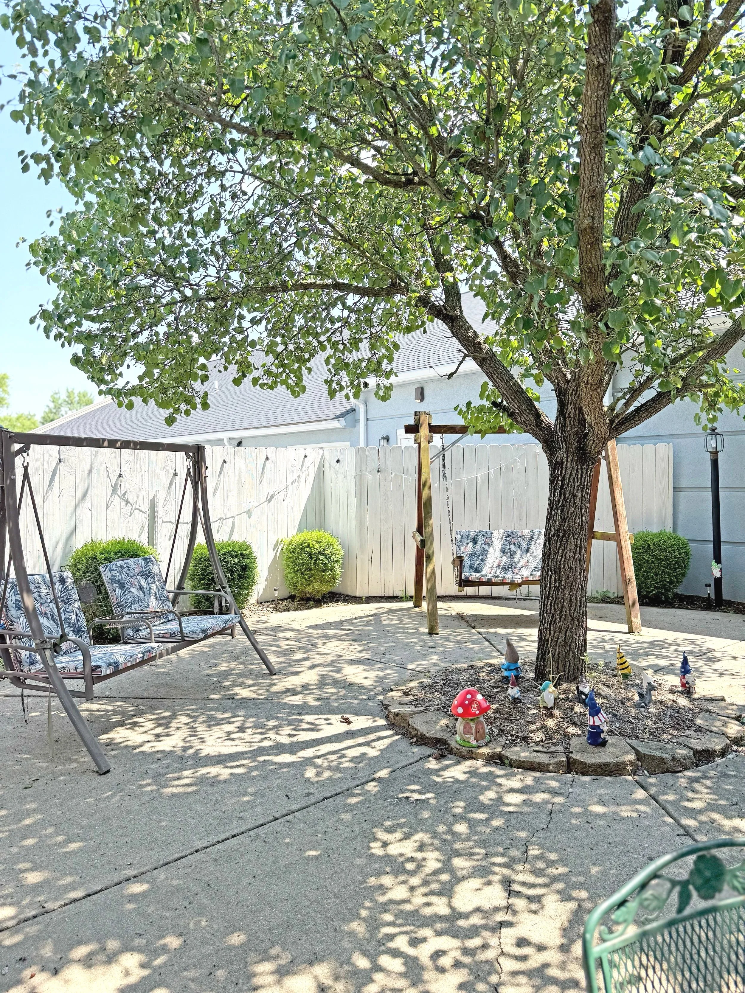 A backyard patio with a large tree, two decorated garden gnome statues, a garden swing, a bench swing, and small bushes, with shadows from the tree on the ground and a white fence in the background.