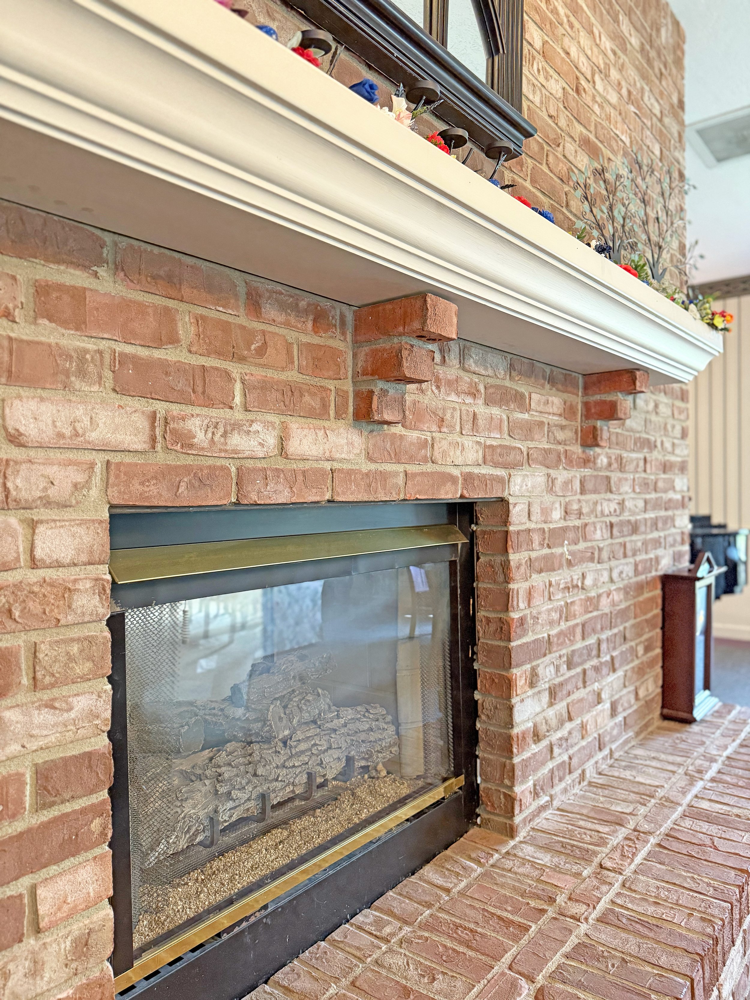 Brick fireplace with glass doors, displaying logs inside, and a white mantle with decorative items above.