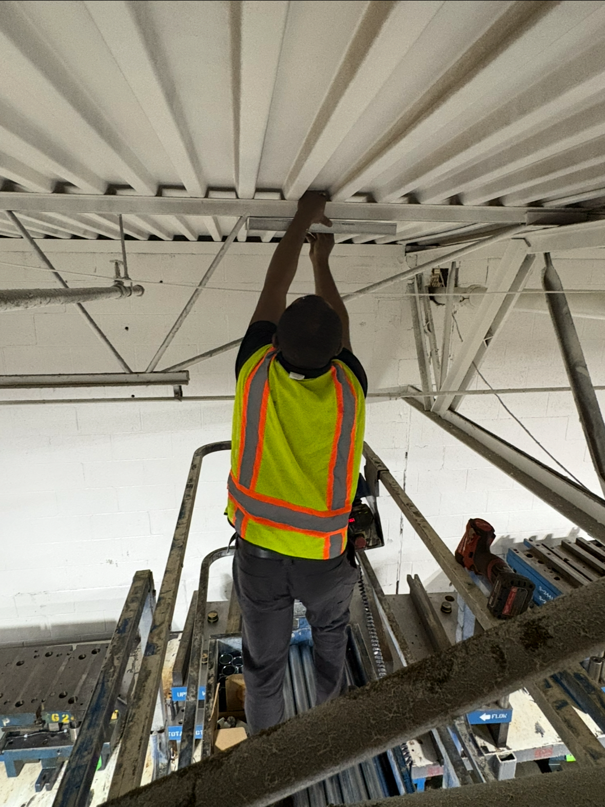 A construction worker in a neon yellow safety vest is installing or repairing ceiling panels on scaffolding inside a building.