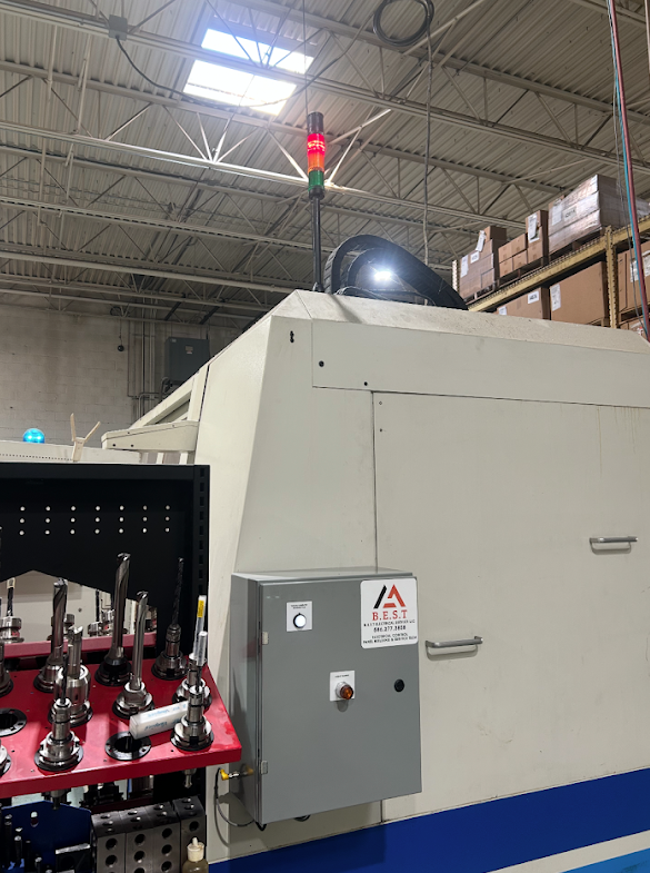 Industrial machinery inside a warehouse with metal beams and boxes on shelves, featuring a control panel with a warning sticker and red and green indicator lights.