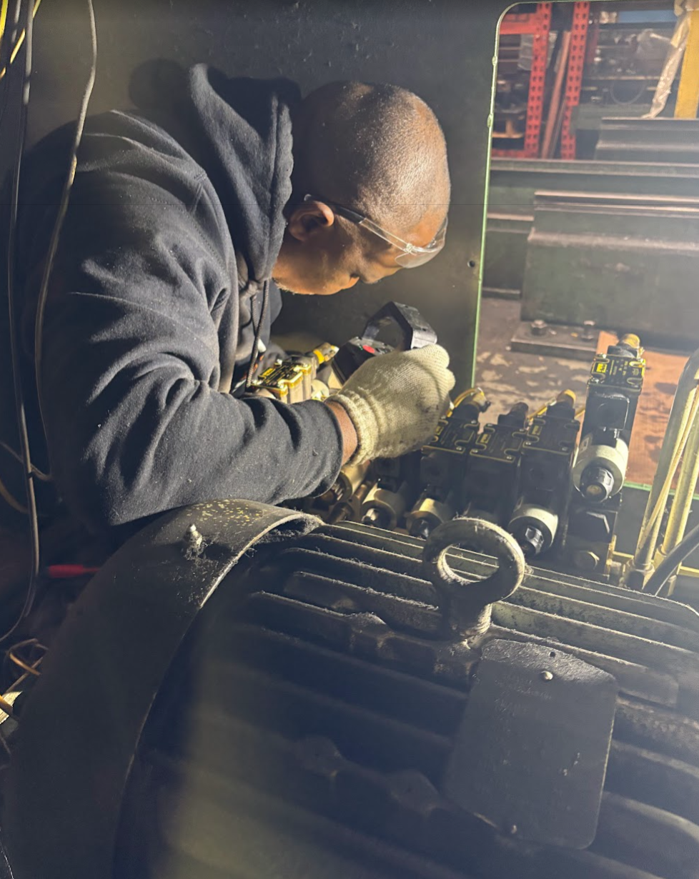 A man working on machinery in a workshop, wearing safety glasses and gloves, with tools and equipment around him.