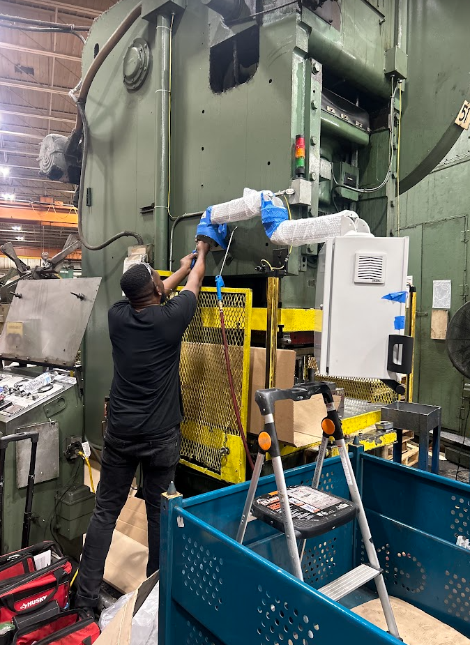 A worker operating industrial equipment in a factory setting, using a ladder to reach the machine.