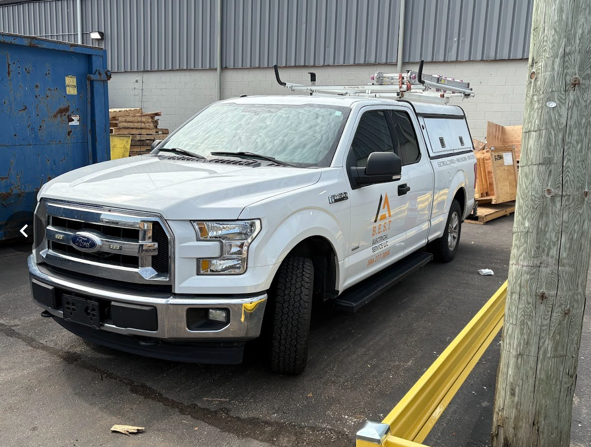 White utility truck with ladder on roof and company logo on door, parked in industrial area with dumpster and wooden pallets nearby.