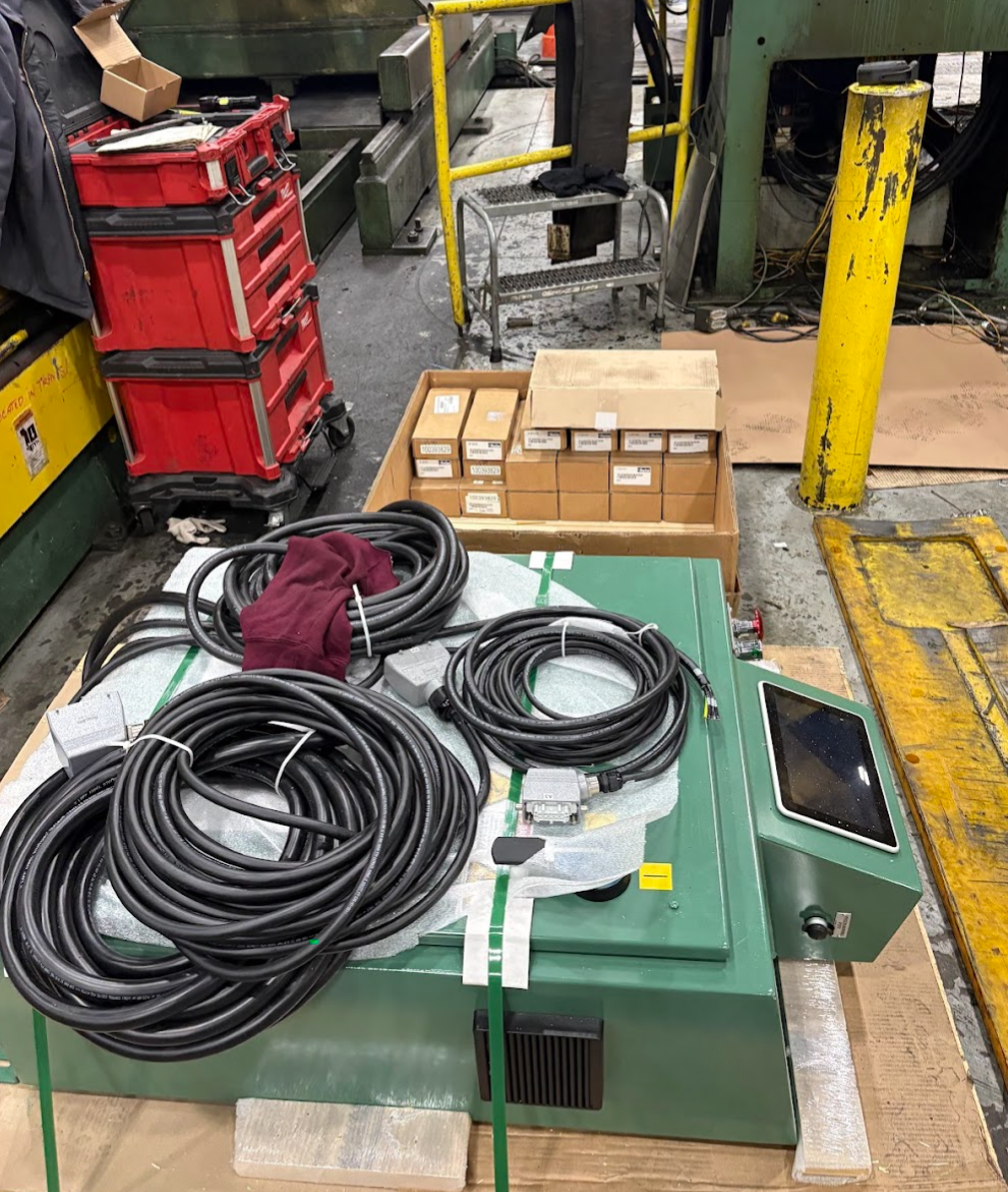 industrial workspace with coiled cables on a green machine, red tool boxes, boxes of parts, and yellow safety barriers.
