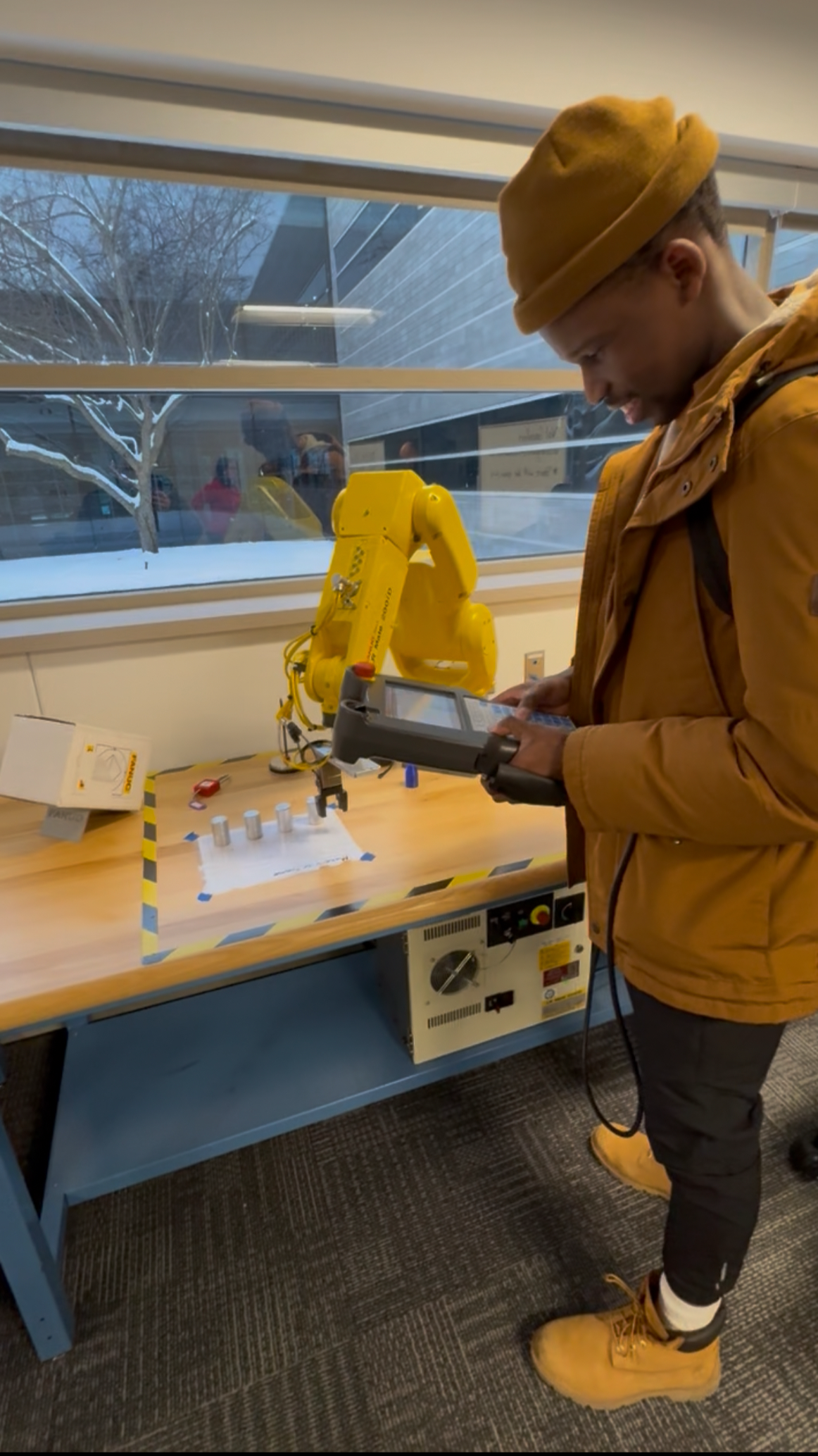 A young man working with a robotic arm in a laboratory, holding a device and standing at a workbench with various tools and components, with a window showing snowy trees outside.