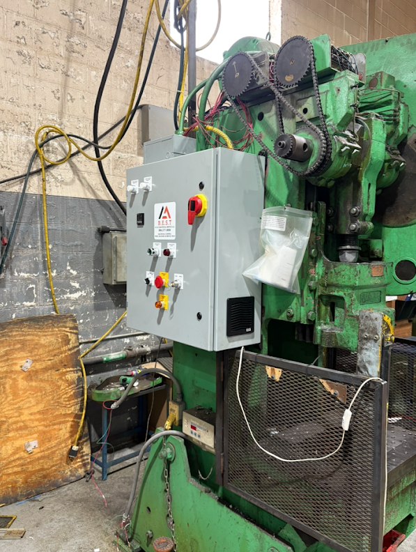 An industrial machine with a gray control panel, emergency stop button, and several interconnected gears and belts in a workshop setting.