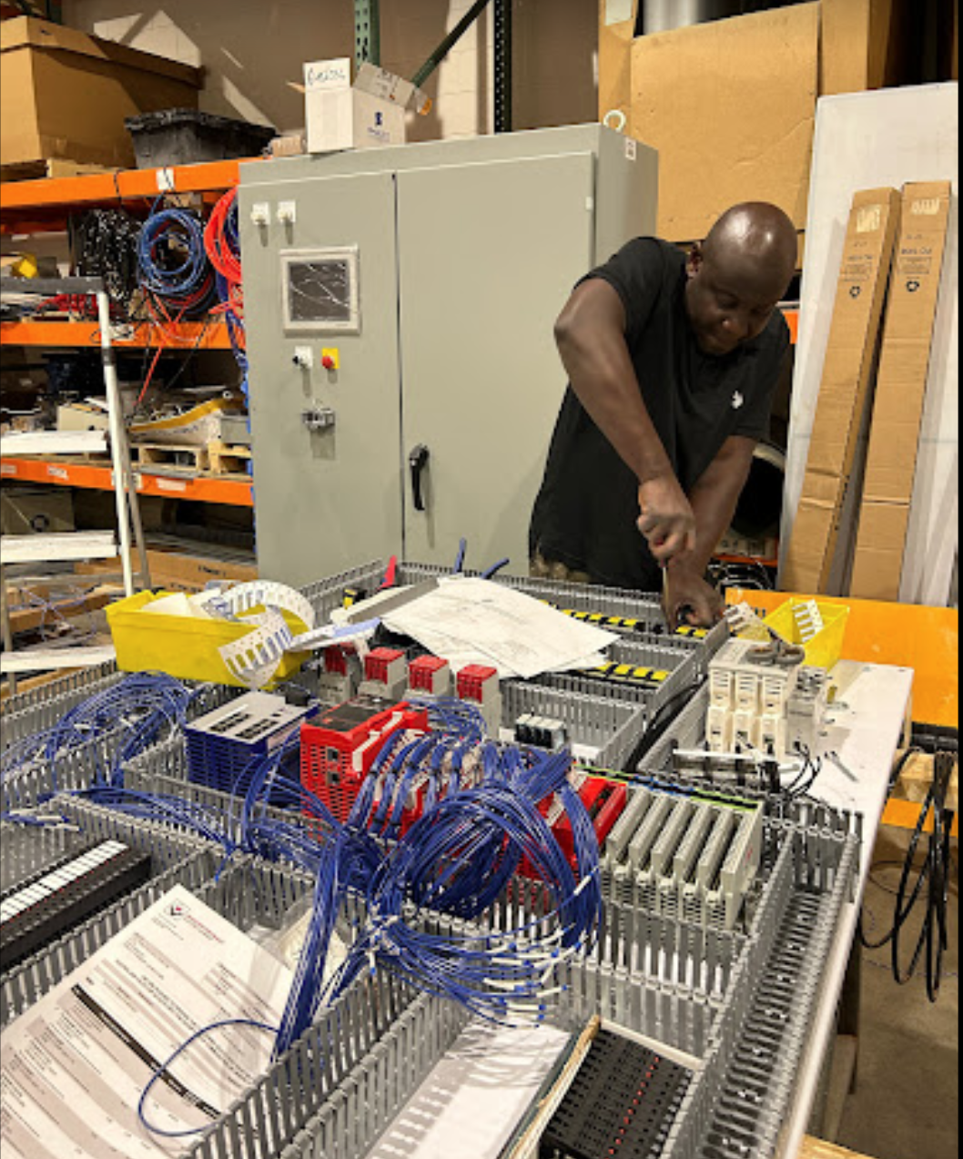 A man working on electrical wiring or control panels at a workbench in a warehouse or workshop filled with components, tools, and storage shelves.