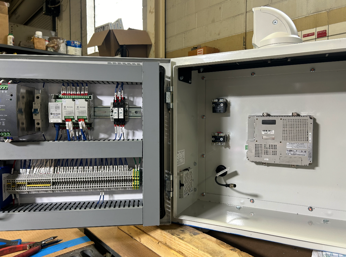 Open electrical control panel with various wires and components inside, situated on a workbench in a workshop.