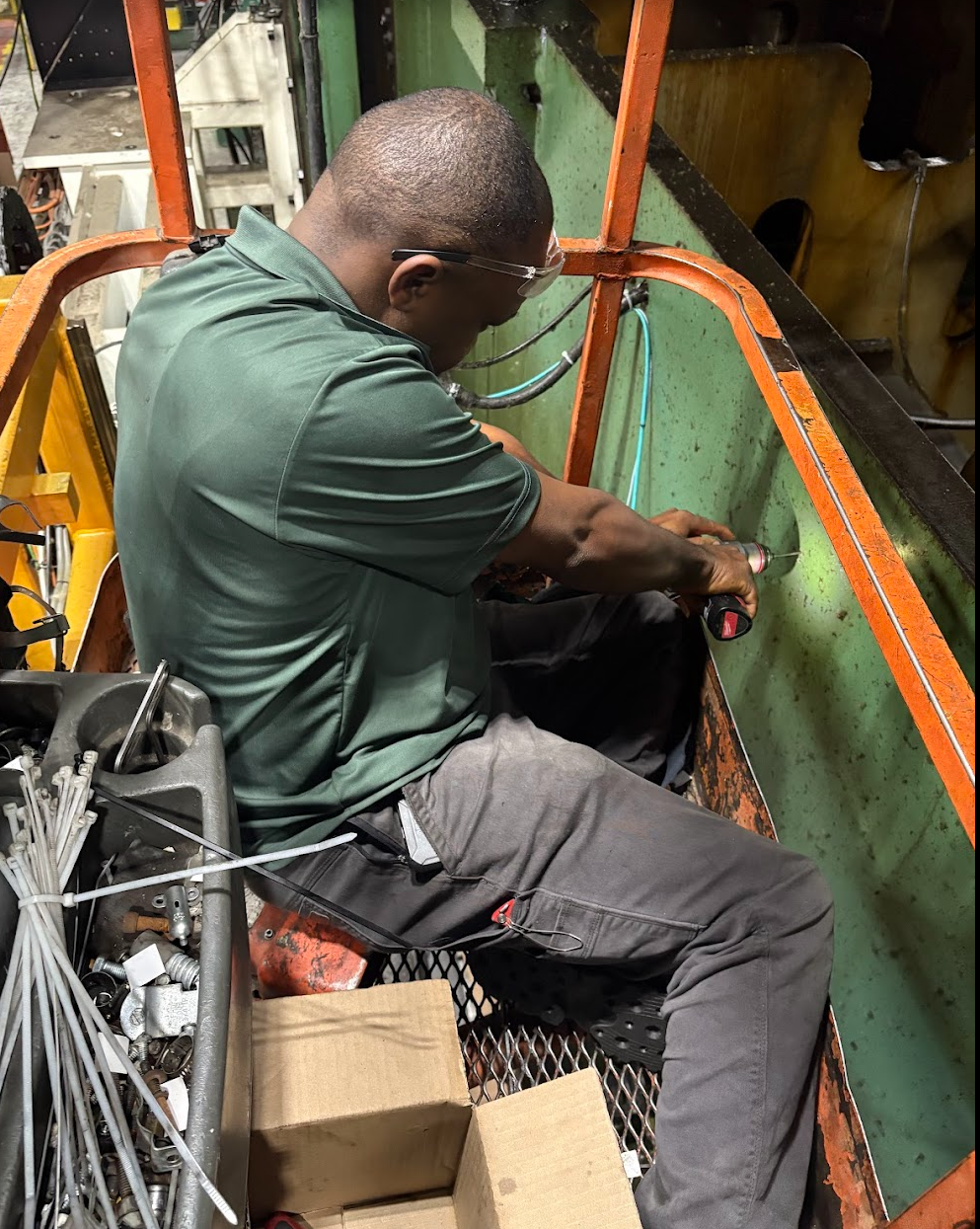 A man wearing safety glasses and a green shirt is working with a power tool inside an industrial or manufacturing setting, surrounded by metal components and equipment.