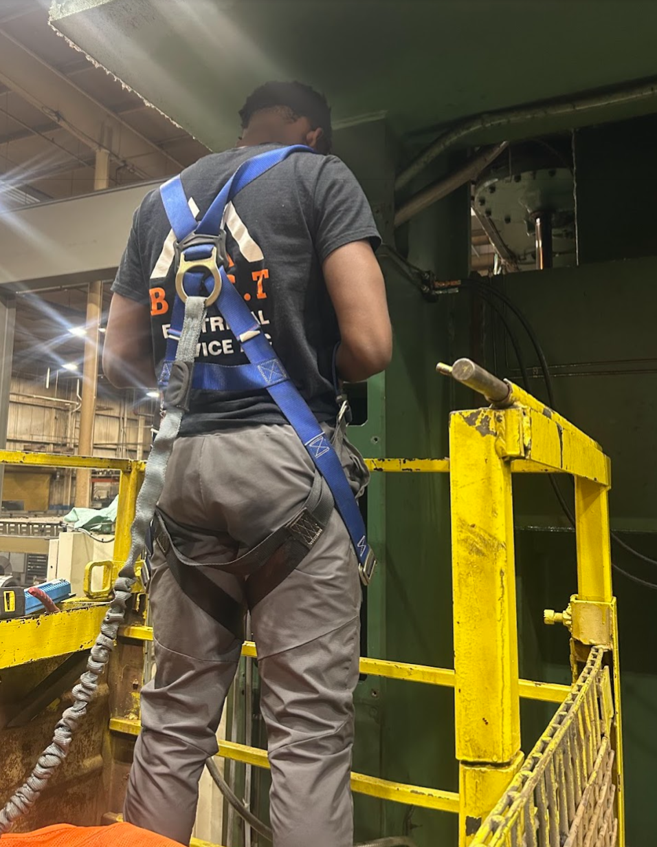 A man wearing work gear with a harness, standing on a yellow safety platform, working on large green equipment in an industrial setting.