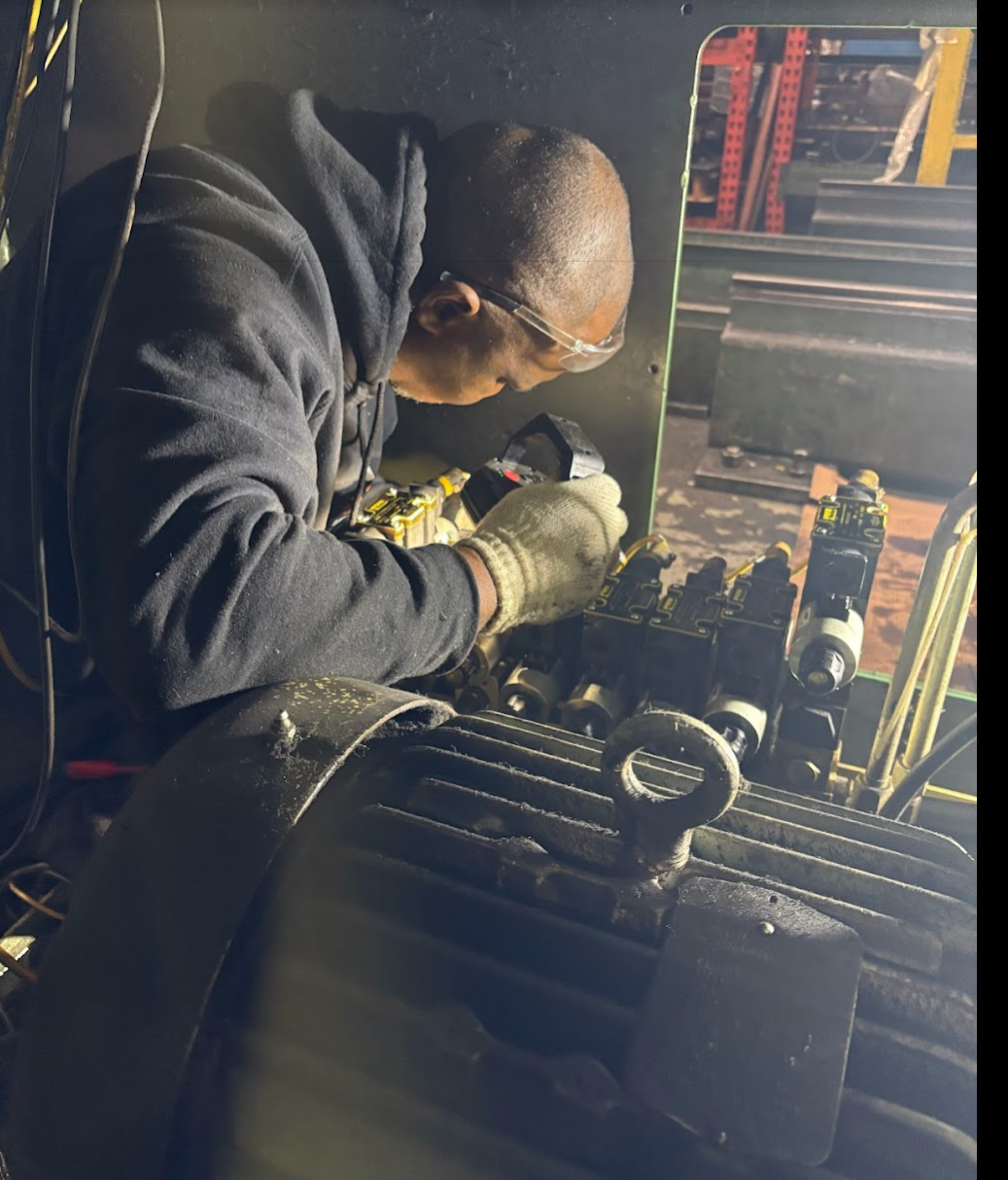 A worker wearing safety glasses and gloves is repairing or assembling machinery in a workshop with metal shelves and tools.