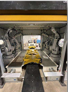 Two robotic arms inside an industrial manufacturing cell working on a yellow component on a conveyor.