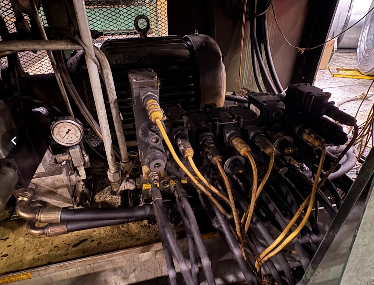 Industrial machinery with multiple hydraulic hoses, a pressure gauge, and electrical components in a workshop or factory setting.
