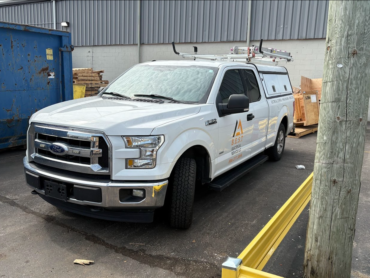 White utility truck parked near a warehouse, marked with electrical service company logo and contact number, with ladder on top and wooden crates nearby.