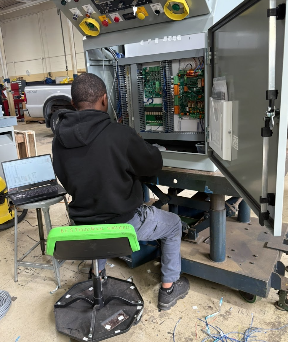 A technician working on an electrical control panel inside a workshop. The person is sitting on a stool with a laptop nearby. The workshop has tools and equipment, including a pickup truck in the background.