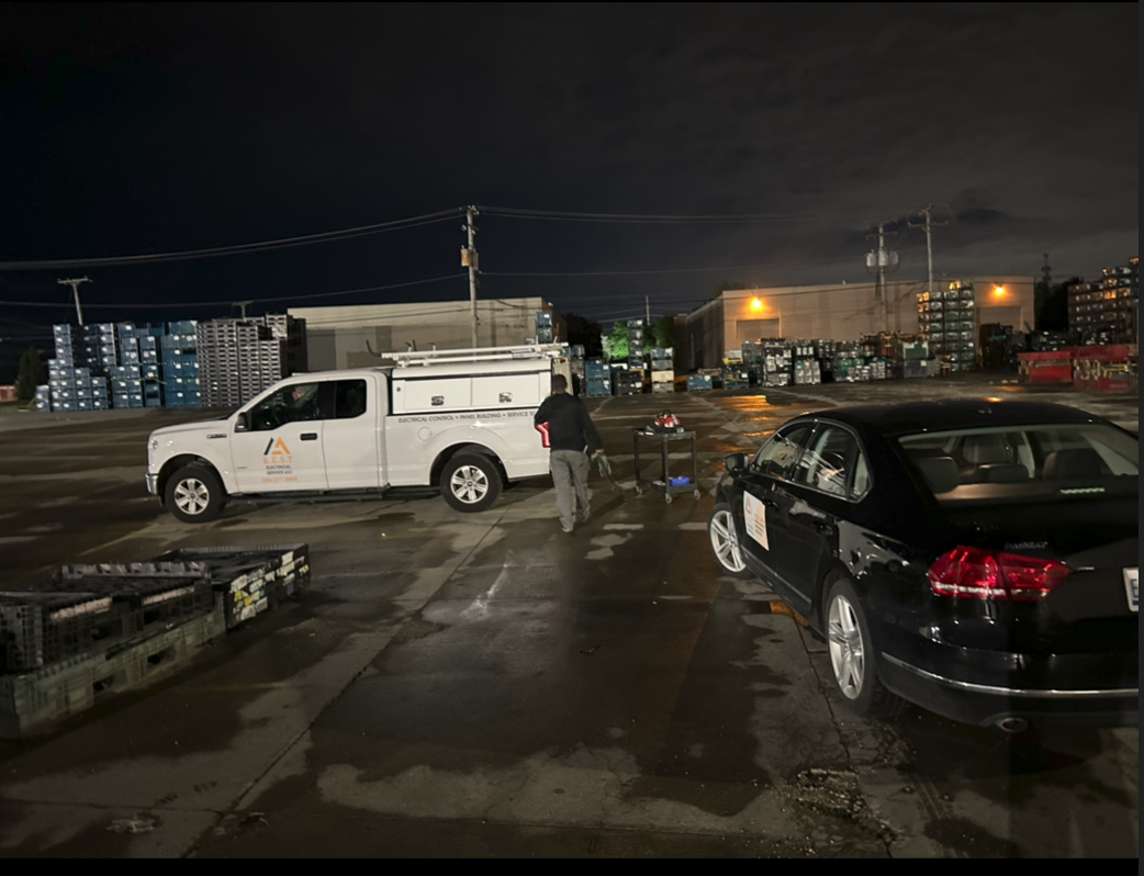 A person standing next to a work truck and a black sedan in a parking lot at night, with storage racks and buildings in the background.