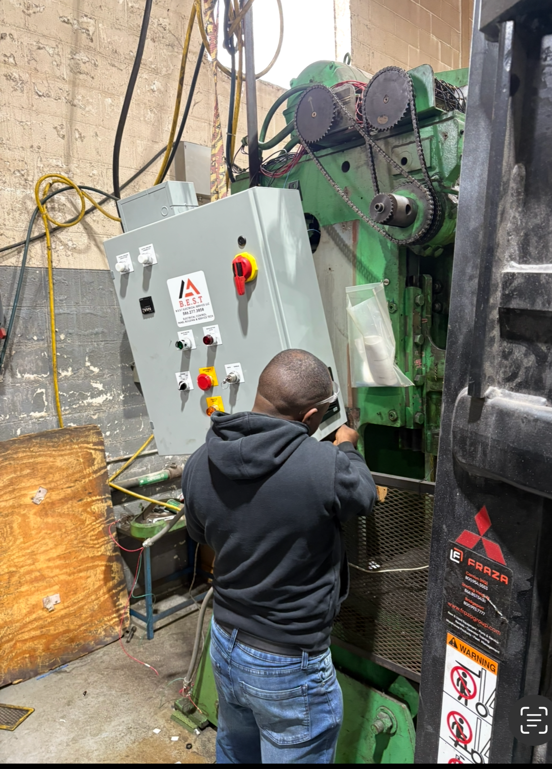 A worker in a black hoodie and jeans is working with a large industrial green machine, adjusting controls on a grey control panel with various switches and buttons inside a workshop with brick walls and electrical wires.