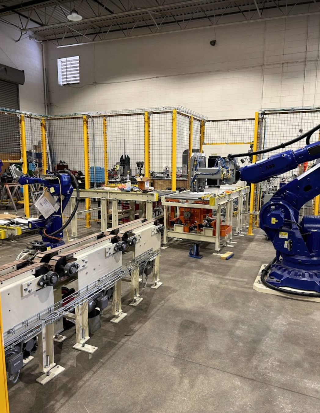 Industrial robotics laboratory with robotic arms, conveyor belts, and machinery, enclosed by safety fencing.