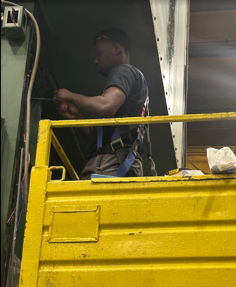 A worker wearing a safety harness and gloves working inside a construction lift with yellow railings in an industrial setting.