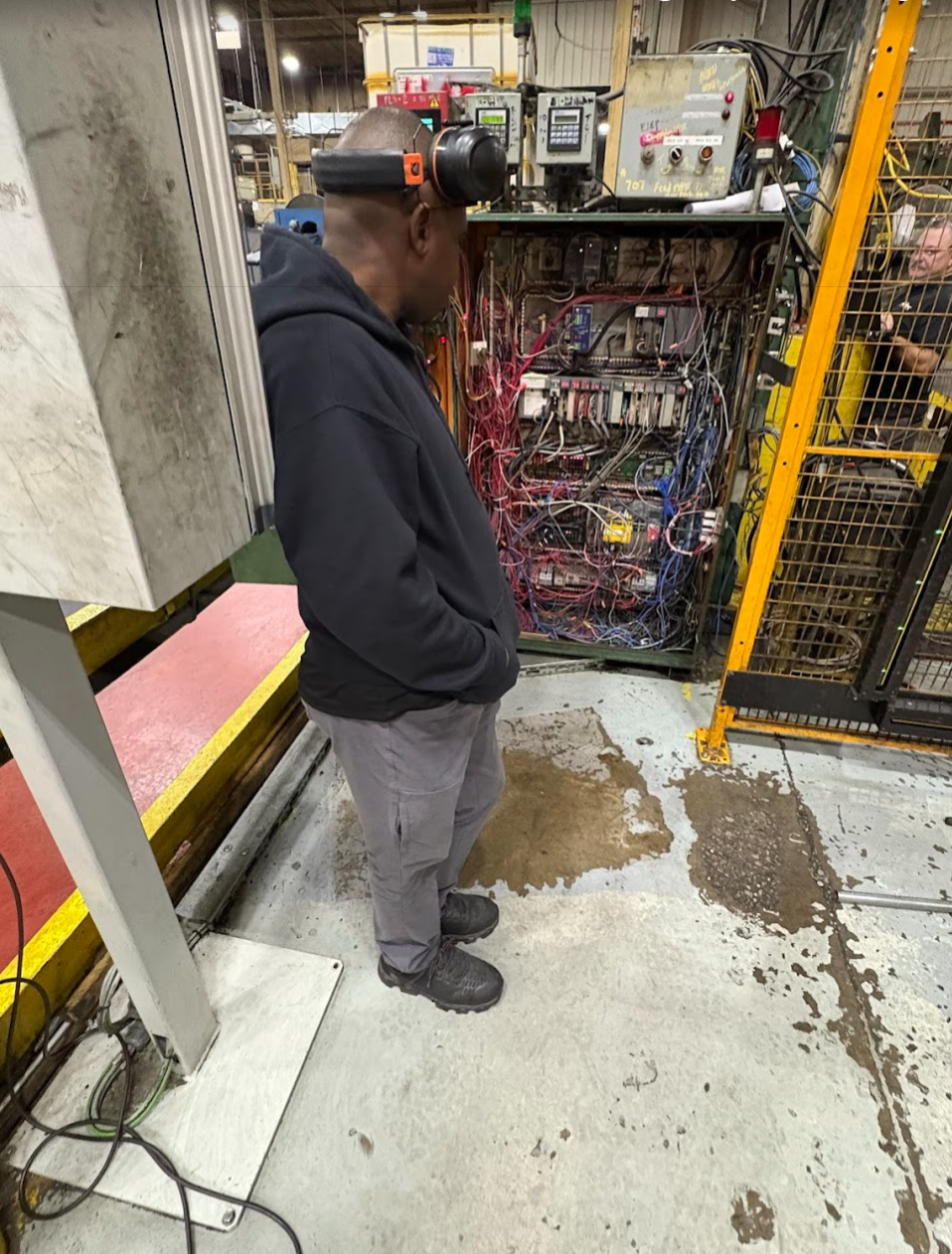 A worker wearing a black hoodie, gray pants, and safety headphones inspects an open industrial electrical panel filled with wires and circuit boards in a factory setting.