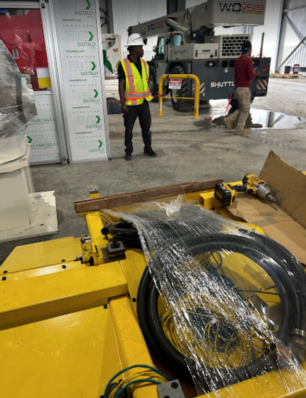 Construction workers in safety vests working in an industrial warehouse with machinery and tools, including a yellow hydraulic lift or crane.