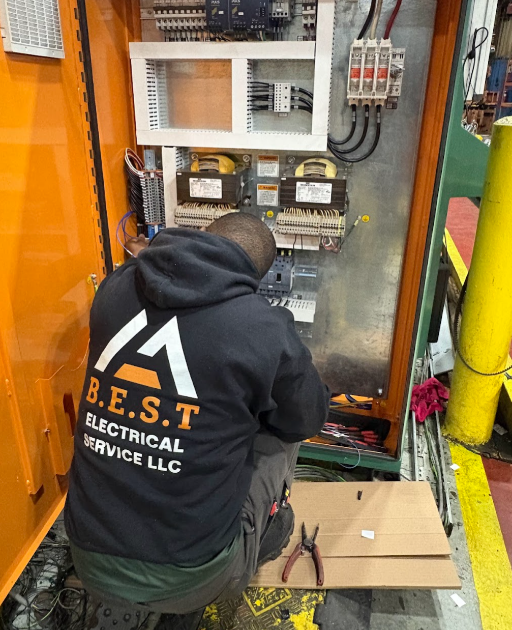 An electrician working inside an electrical control panel, with tools and wiring around him, wearing a jacket labeled 'B.E.S.T Electrical Service LLC'.