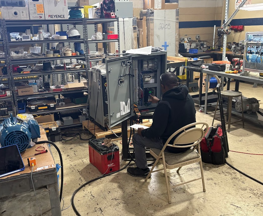 A person working on an open electrical control panel in a workshop cluttered with tools and equipment.