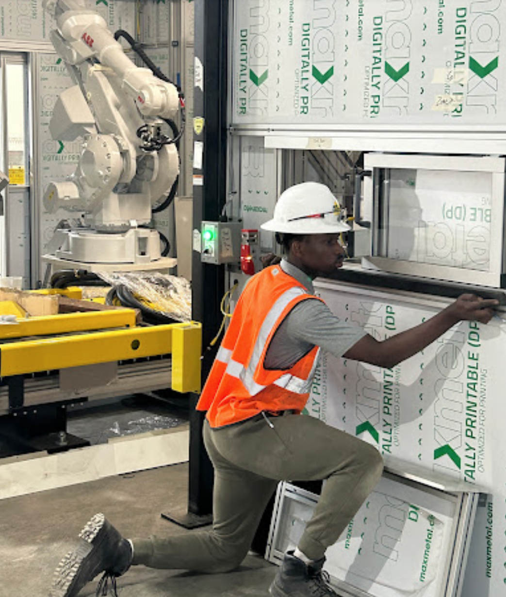 A construction worker wearing a white hard hat and orange safety vest kneeling on one knee, installing or inspecting a large insulated glass window panel at an industrial construction site.