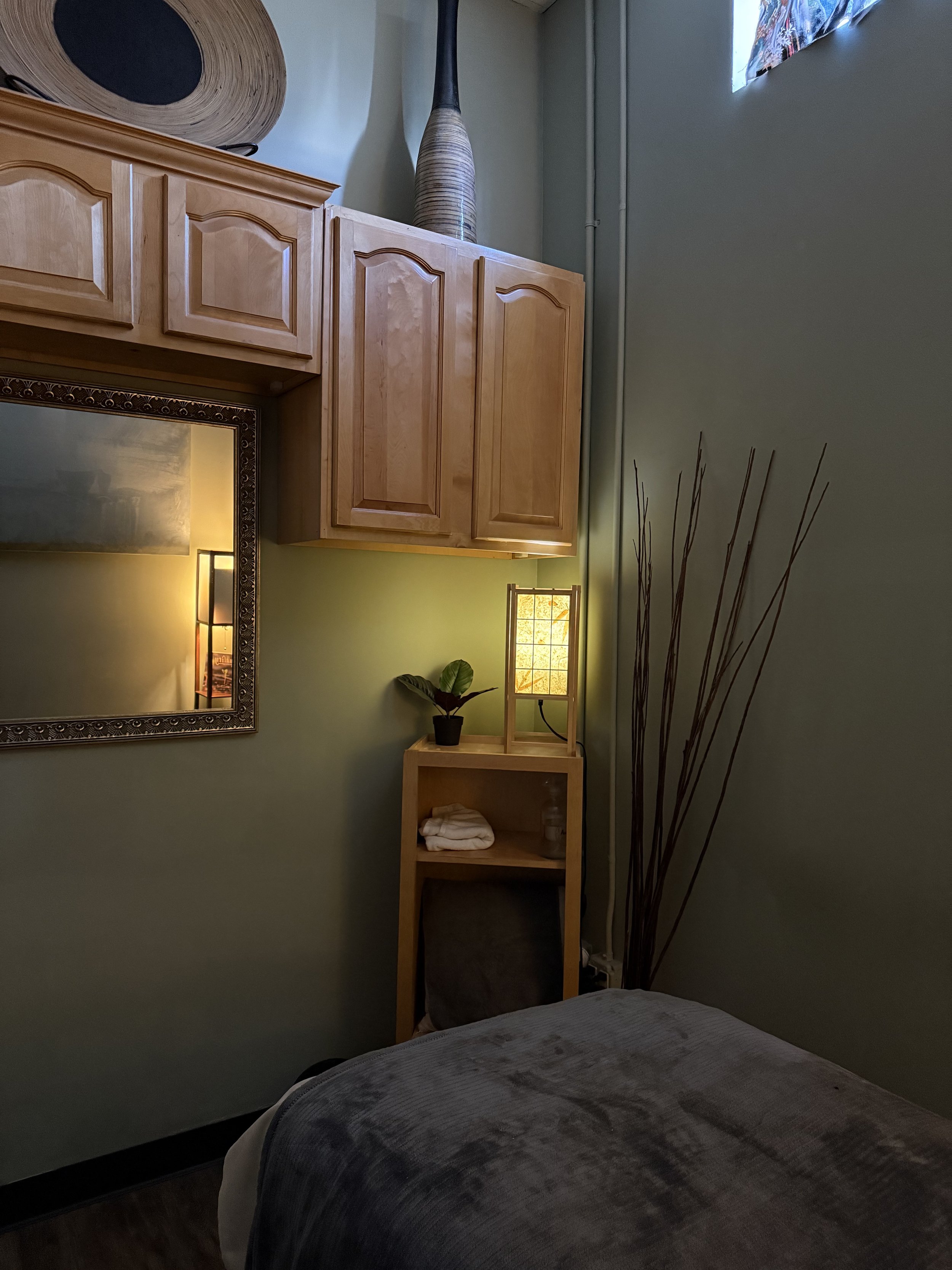 A cozy bedroom corner with wooden cabinets, a mirror, a small wooden shelf with a potted plant and folded towels, a lamp, and tall decorative sticks.