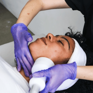 A woman receiving a facial treatment from a professional wearing purple gloves, lying down with a white towel wrapped around her head.