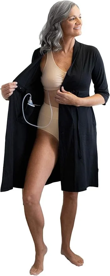 An older woman with gray hair smiling, wearing a black robe over beige underwear, holding open her robe to reveal a dialysis access port on her abdomen connected to a dialysis machine.