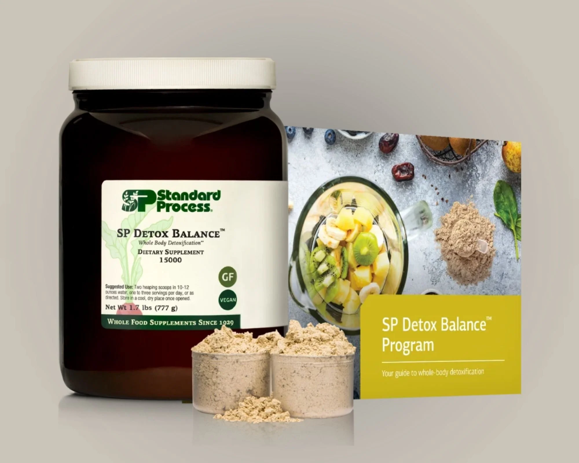 A bottle of Standard Process SP Detox Balance supplement, two scoop-sized containers of powder, and an informational brochure about the detox program, with a background of a fruit smoothie and various natural ingredients.