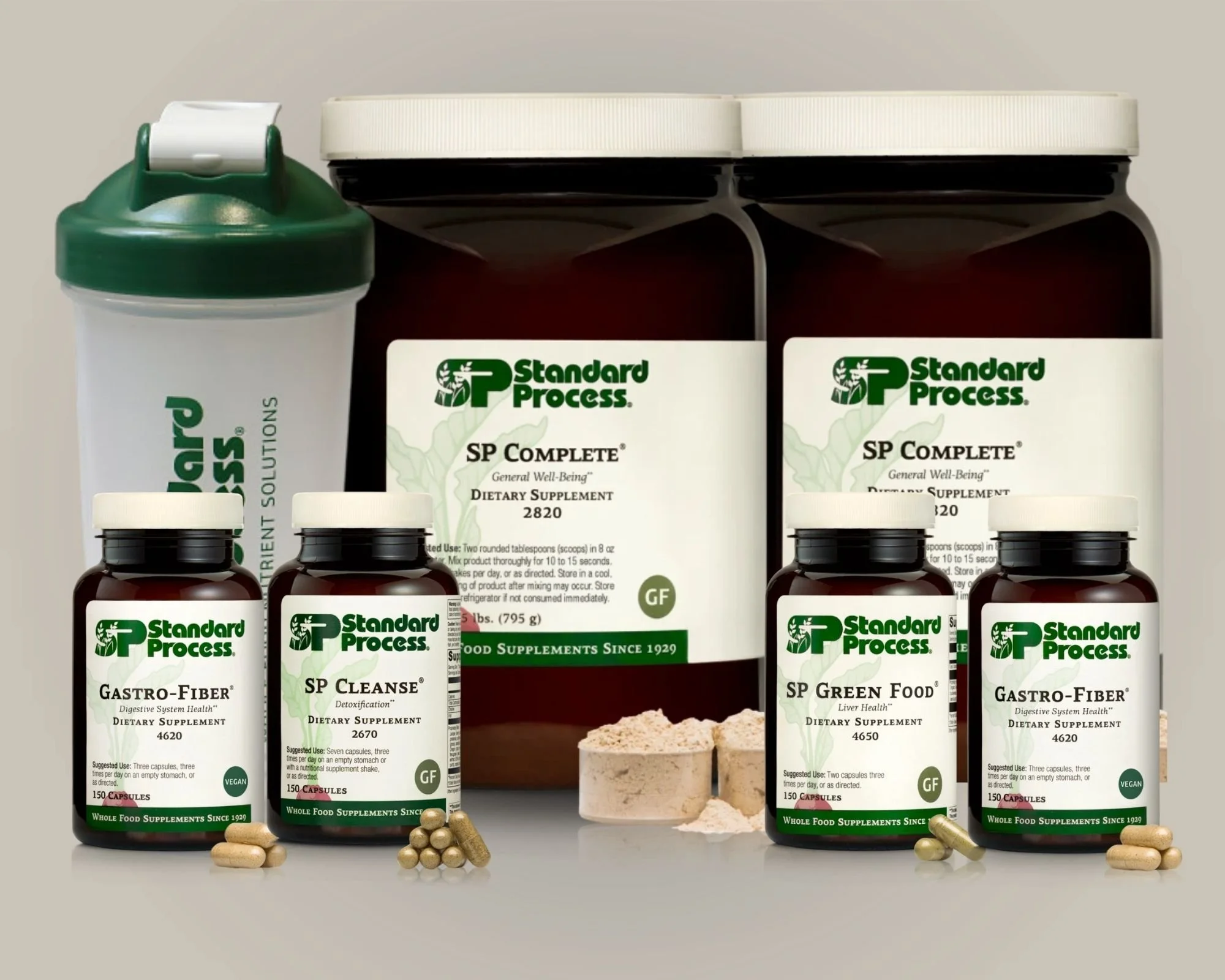 Collection of dietary supplement bottles and containers with white lids, featuring labels from Standard Process. The products include SP Complete, Gastro-Fiber, SP Cleanse, and SP Green Food, along with a shaker bottle and some powdered supplement.