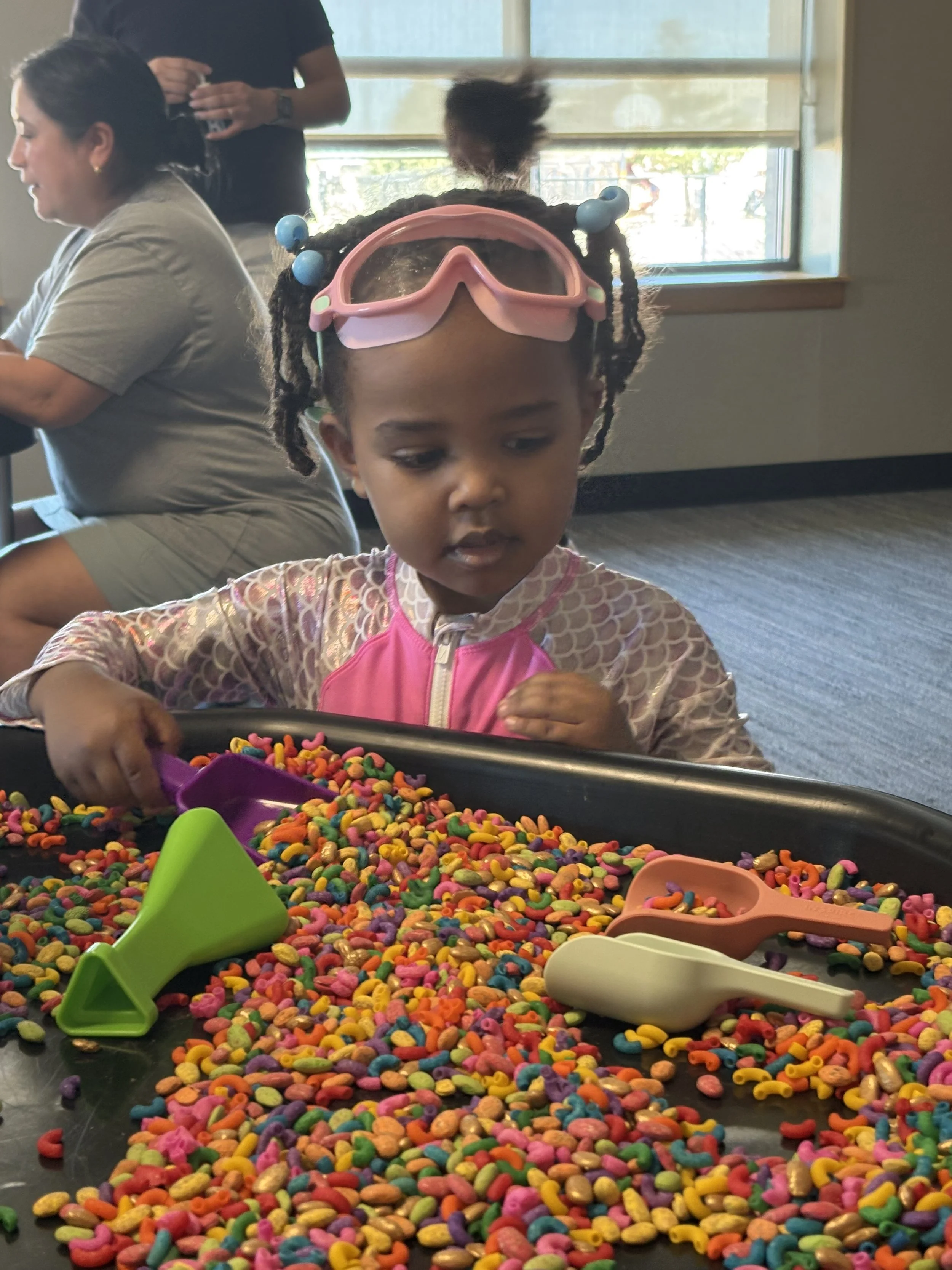 A young girl with braided hair and pink goggles on her head is playing with colorful cereal pieces on a black tray, using various scoops, in an indoor setting with large windows in the background.