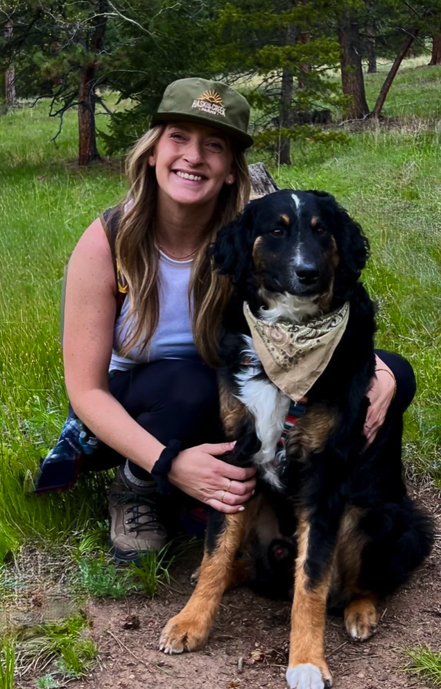 A woman with long blonde hair, smiling, kneeling outdoors next to a large black and tan dog with a bandana around its neck, surrounded by green grass and trees.