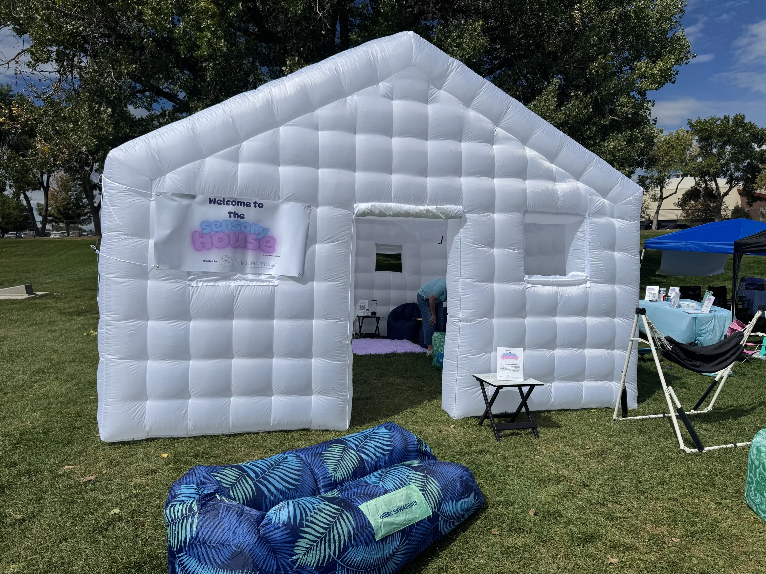 An inflatable white house tent with a sign reading "Welcome to the Sensory House" inside an outdoor park. There are trees, a blue sky, and other tents and chairs nearby.
