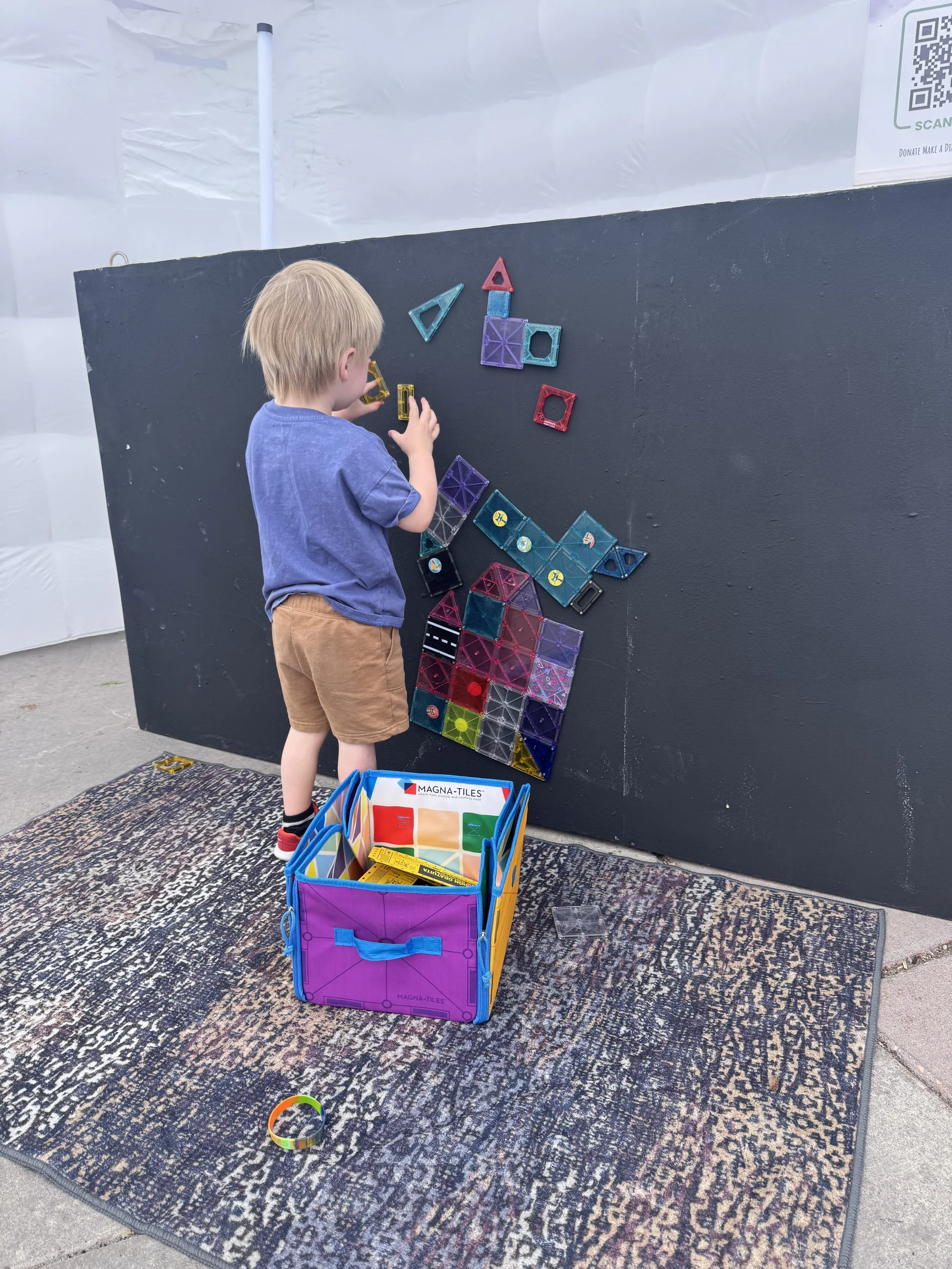 A young boy building with colorful magnetic tiles on a black wall at an outdoor event.
