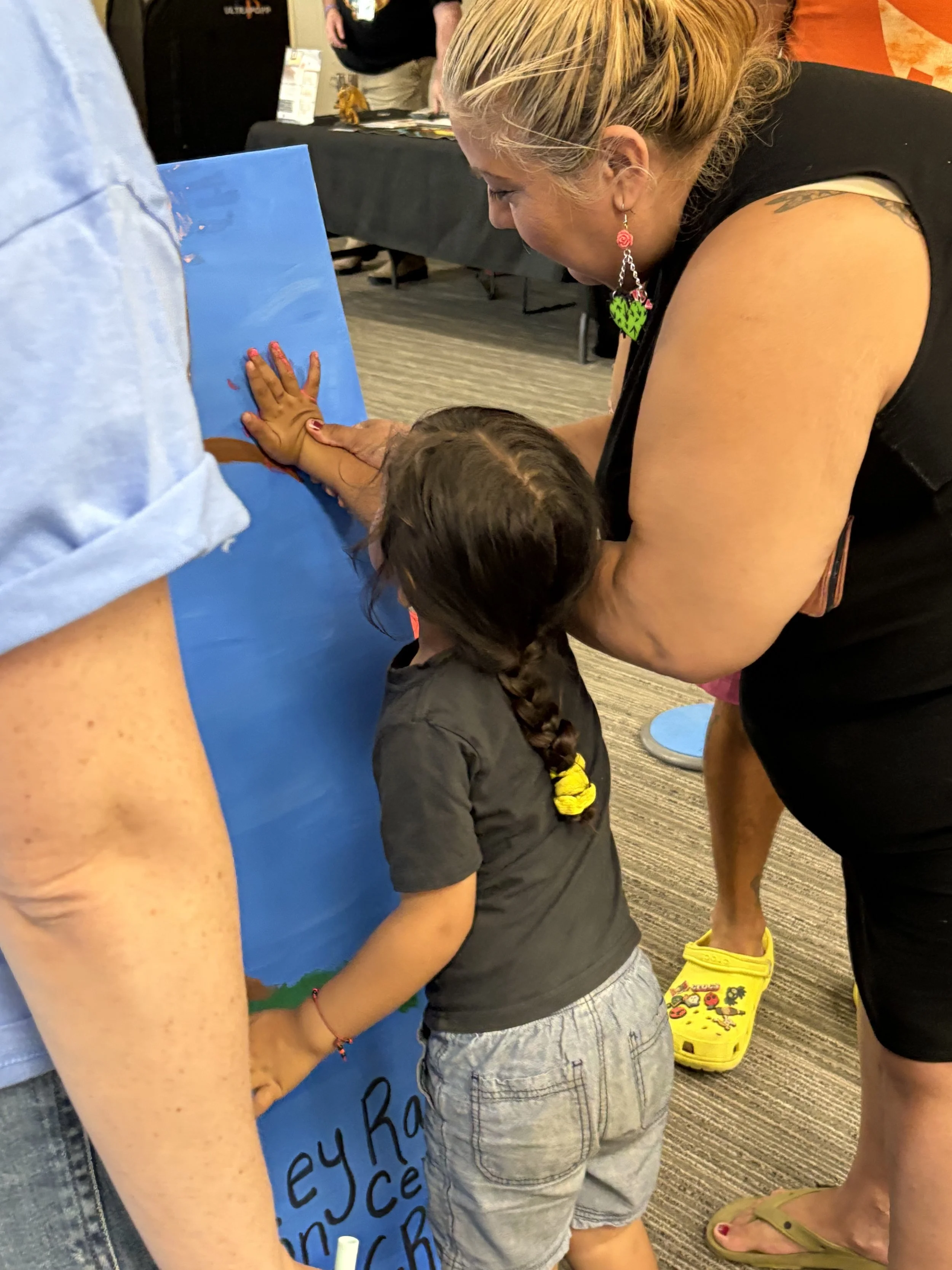 A woman and a young girl are painting a mural together on a large blue poster board. The woman is leaning over, guiding the girl who has braided hair and is wearing a dark t-shirt and shorts. The girl's hand is covered in red paint as they both work on painting a tree or similar scene. Other people and tables are visible in the background.