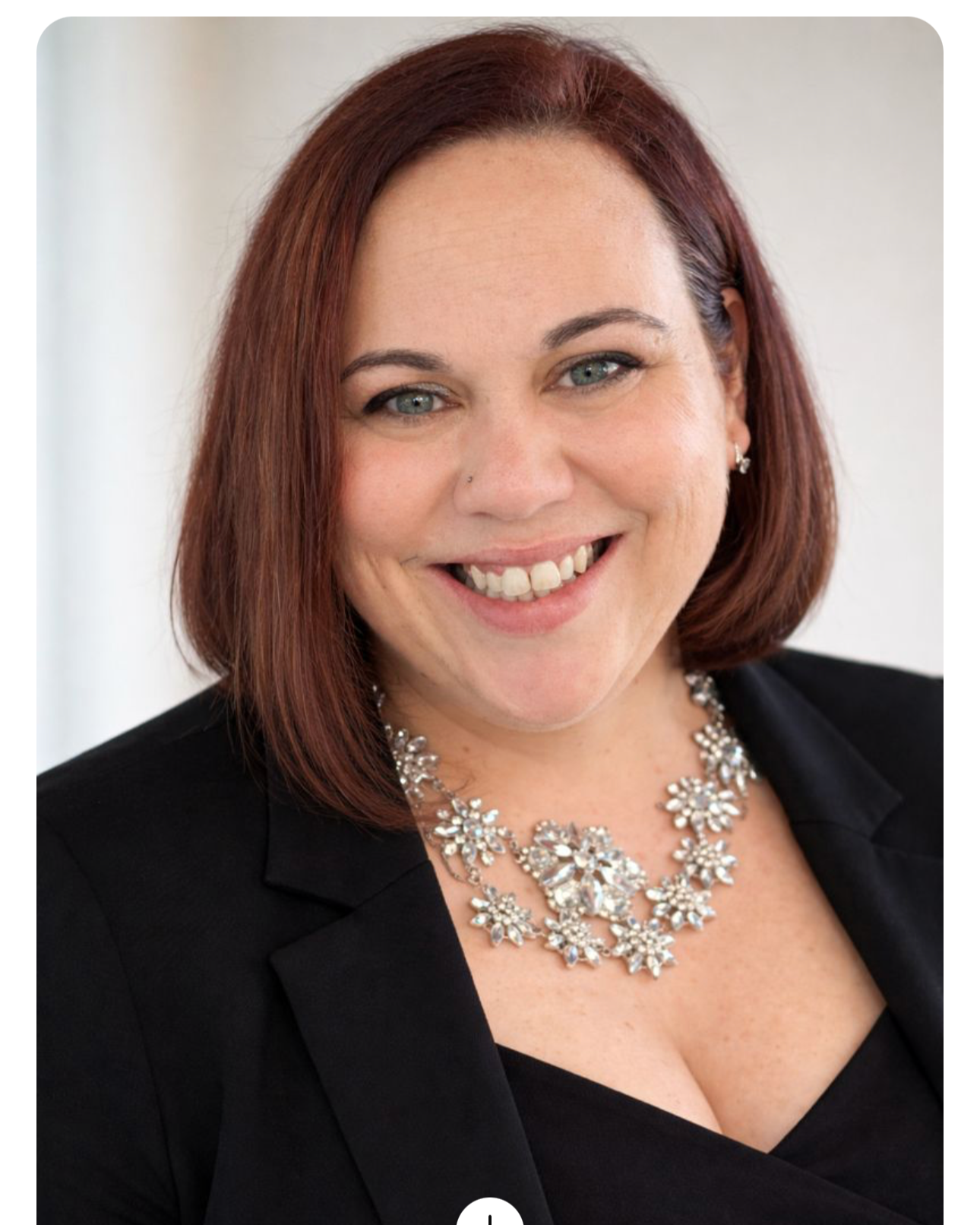 A woman with red hair, wearing a black blazer, and a sparkling floral necklace, smiling at the camera.