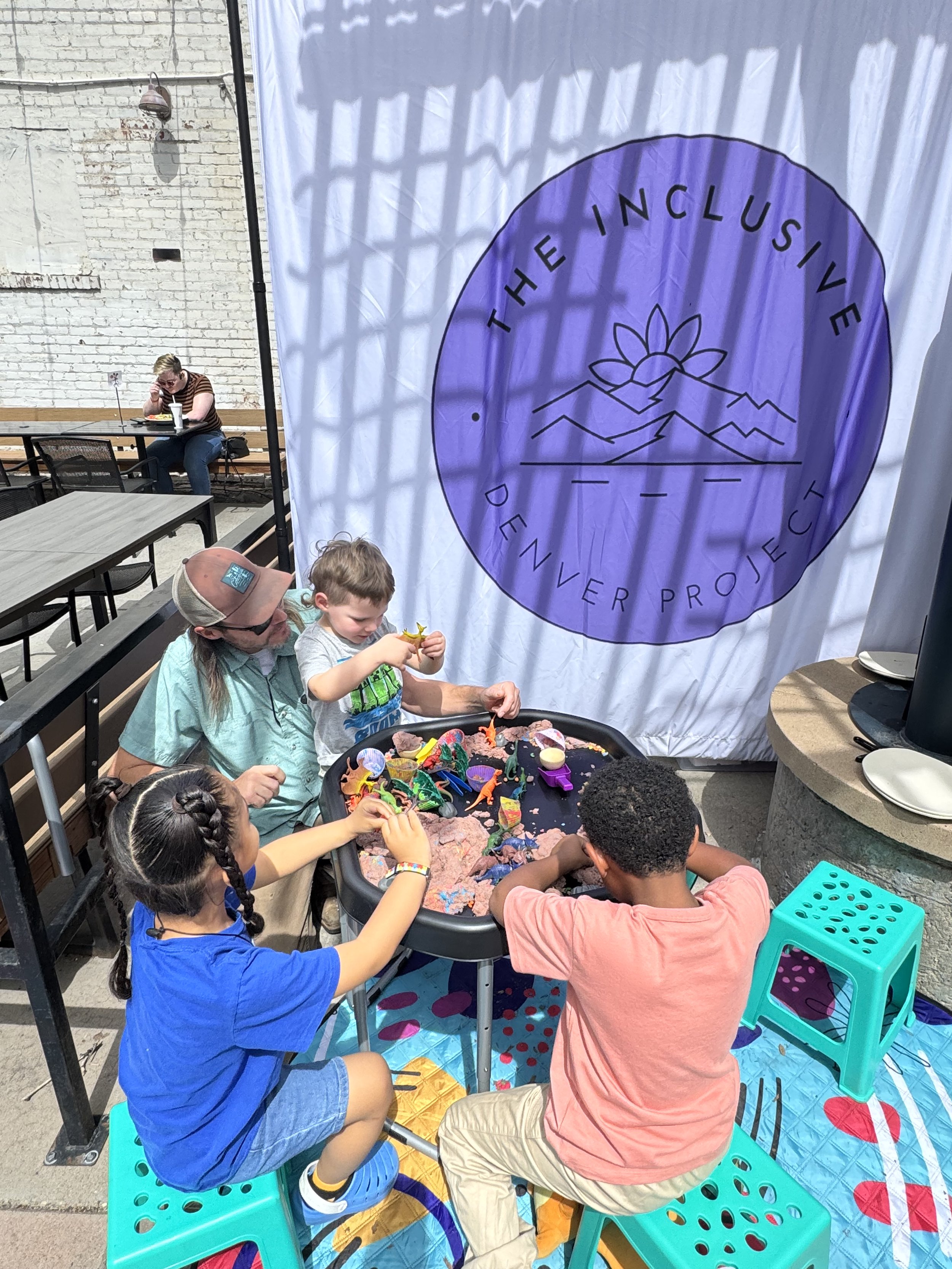 Children playing with toys at a sensory table outdoors, with a large banner reading 'The Inclusive Denver Project' and a woman sitting at a nearby table.