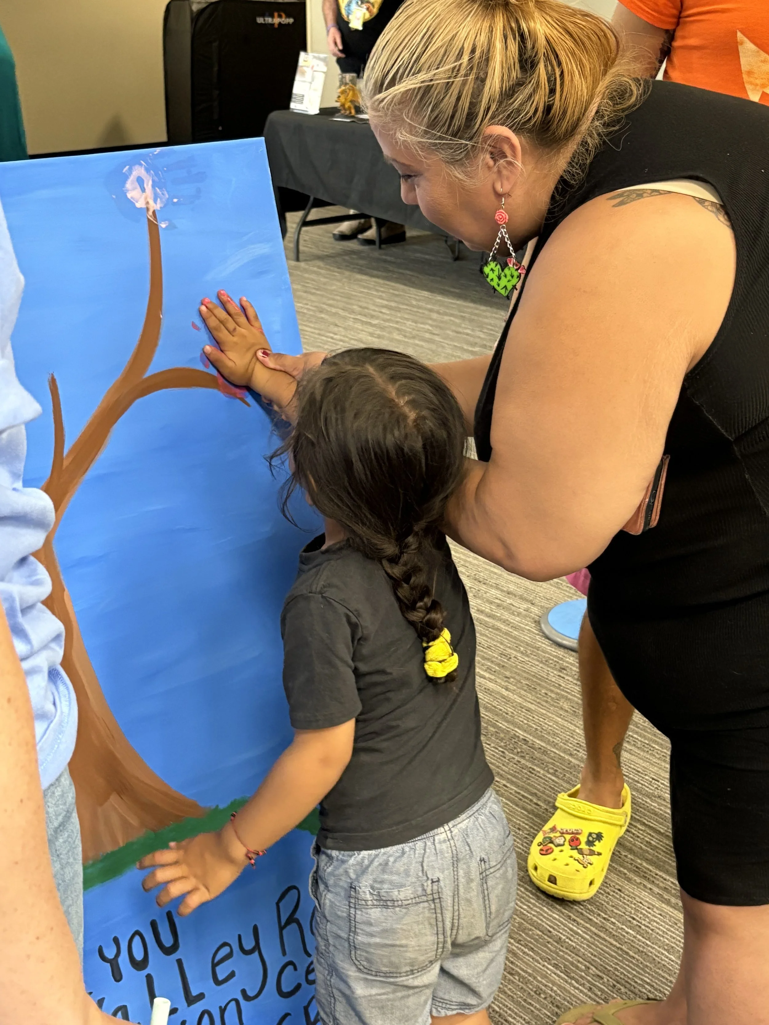 A woman with blonde hair and earrings with a child with a braid learns about a painted tree on a blue background at a table in a room with a carpeted floor.