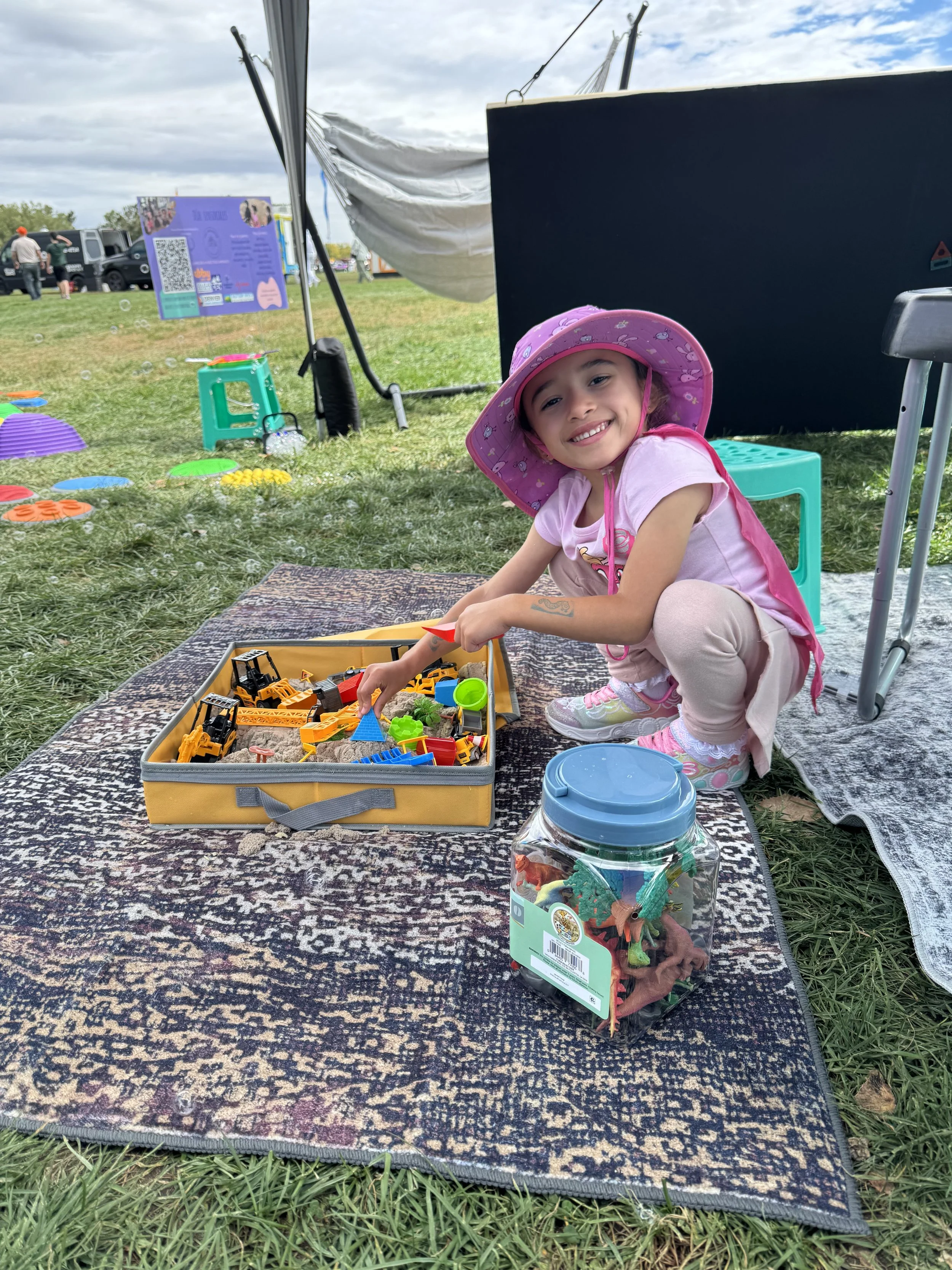 A young girl wearing a pink hat and pink clothes smiling and playing with toy construction vehicles on a patterned rug outdoors at a park or fair, with a jar of colorful dinosaur toys nearby and houses or tents in the background.