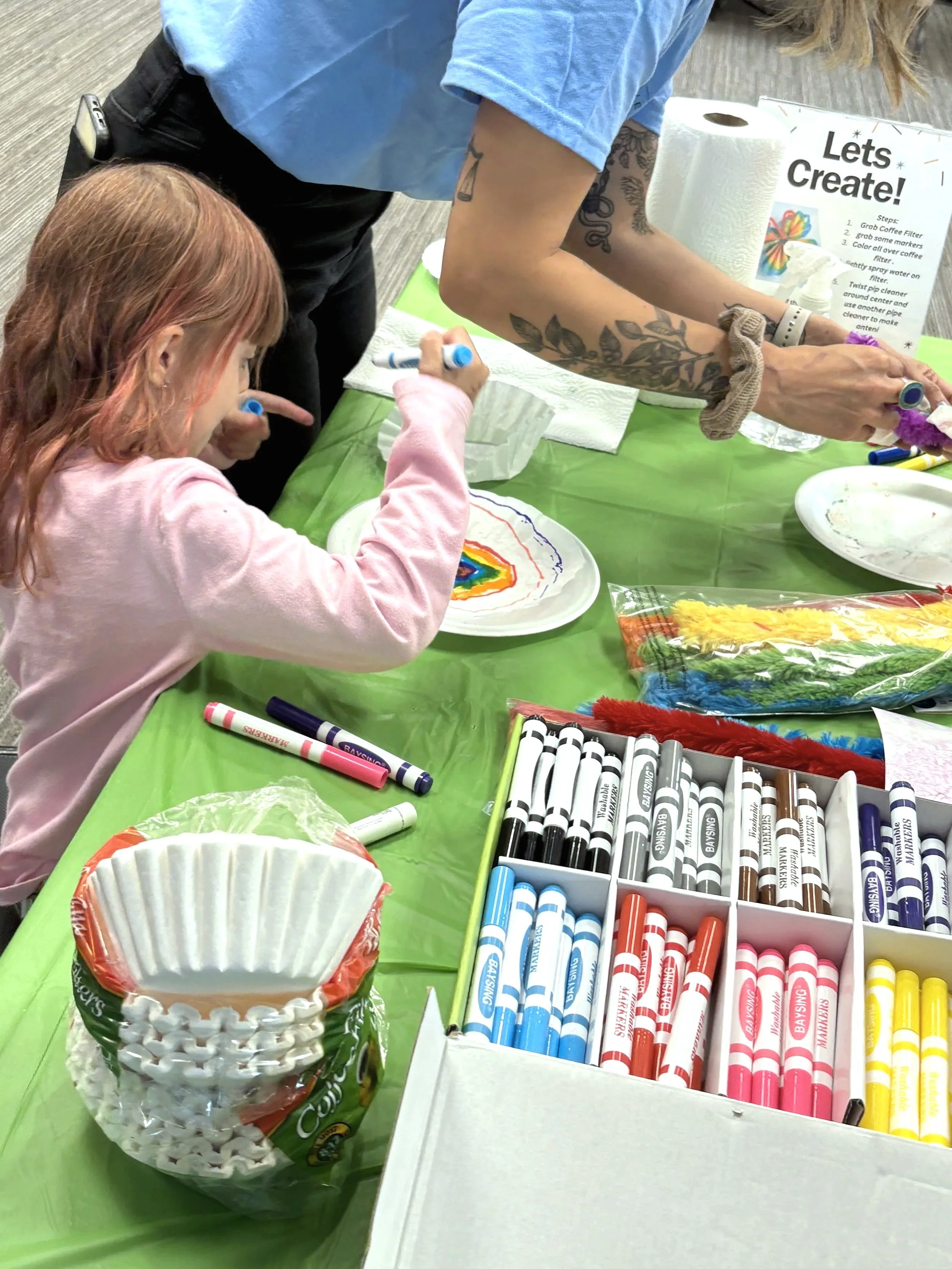 A young girl and an adult are decorating ceramic plates with colorful markers at a craft table covered with a green tablecloth. The table has markers, a package of paper cups, and decorative supplies. There is a sign in the background titled 'Let's Create!'