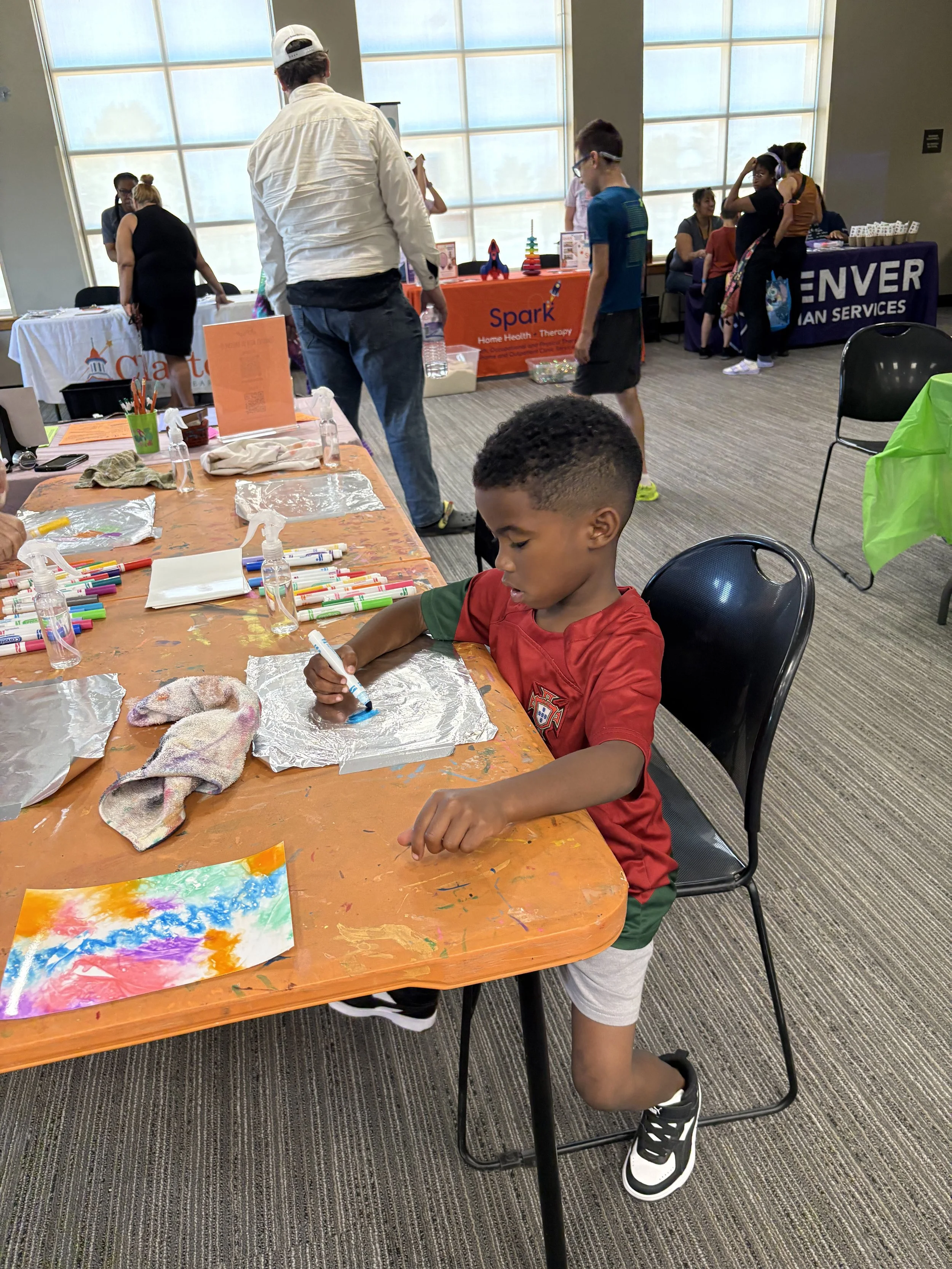 A young boy wearing a red soccer jersey and gray shorts is sitting at a table, drawing on a piece of aluminum foil with a blue marker. The table is covered with various art supplies, including colored markers, spray bottles, and cloths. In the background, other children and adults are browsing booths at an indoor event with large windows letting in natural light.