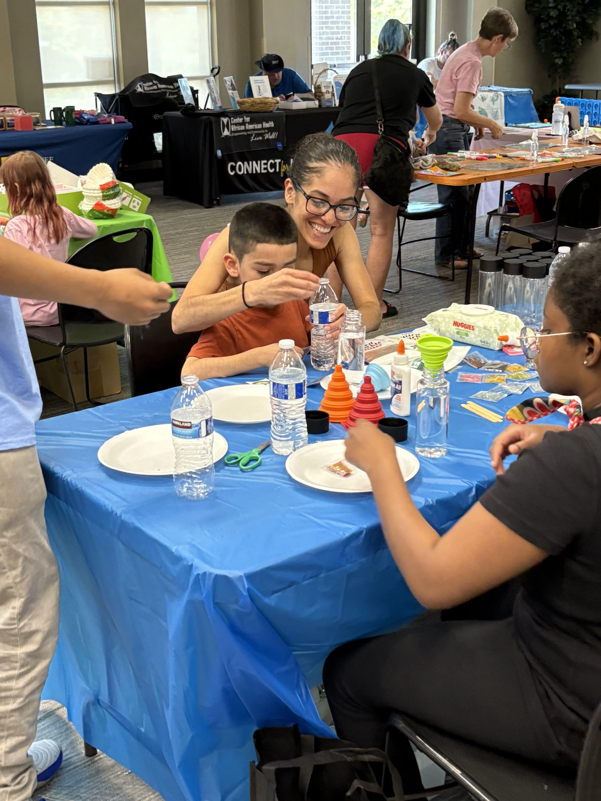 A group of children and adults sitting at a table with craft supplies, playing a game with orange and red cones, in a room with tables, chairs, and informational booths in the background.
