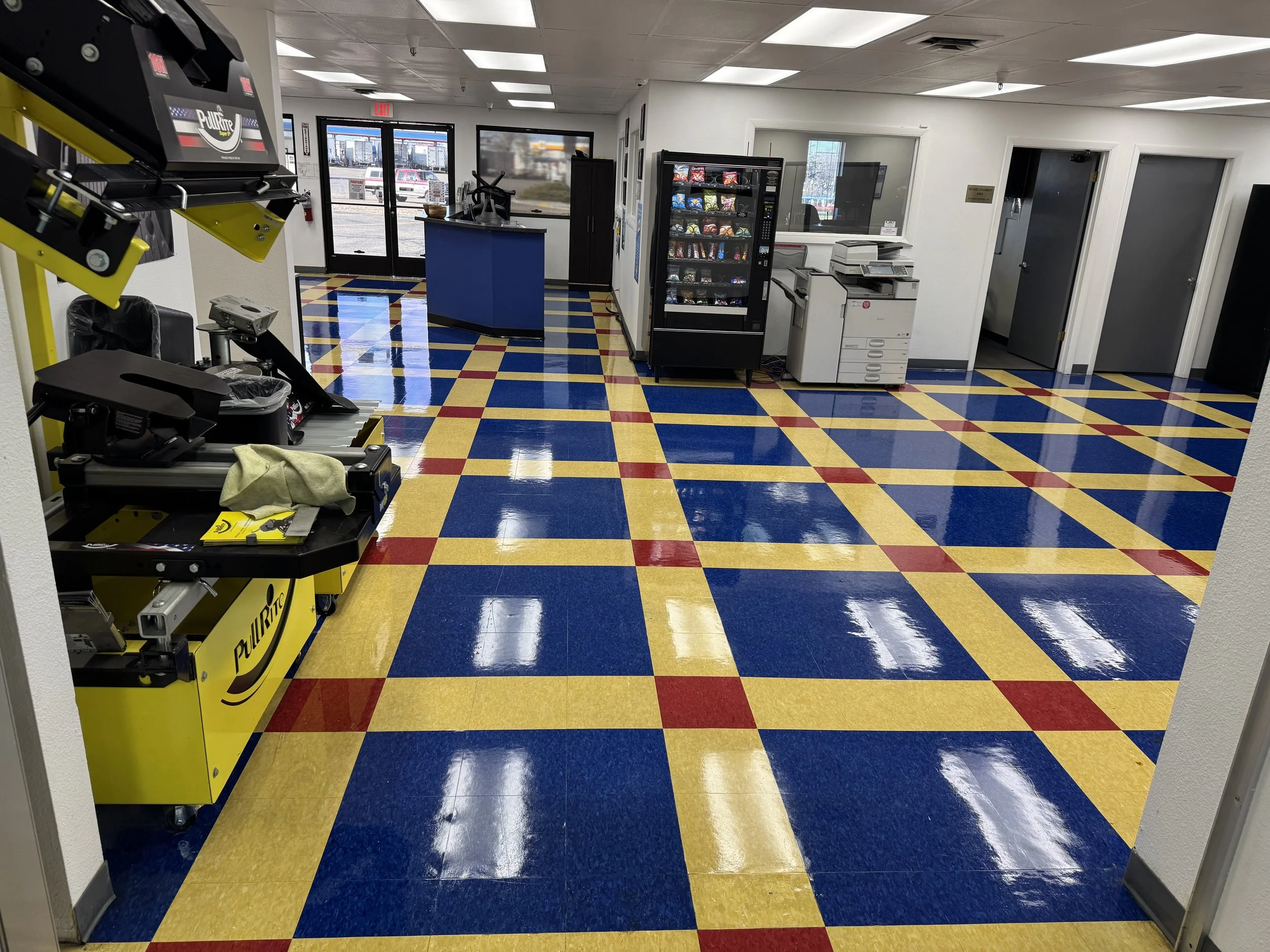 Interpreted view of a convenience store or small retail shop with a checkered yellow, red, and blue tiled floor, vending machines, and a checkout counter near the front door.