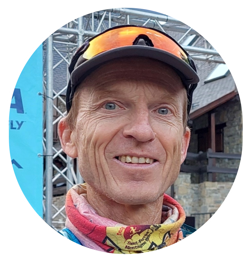 A smiling man wearing sunglasses, a helmet, and a colorful neck gaiter, standing outdoors in front of a metal structure and building, likely at a sporting event or outdoor activity.