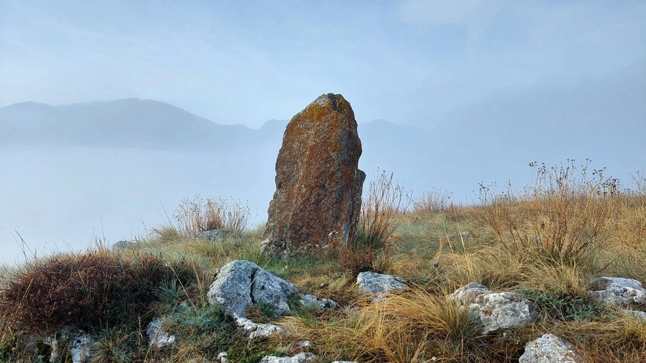 A menhir stands on a grassy knoll against a backdrop of misty Pyrenean mountains.
